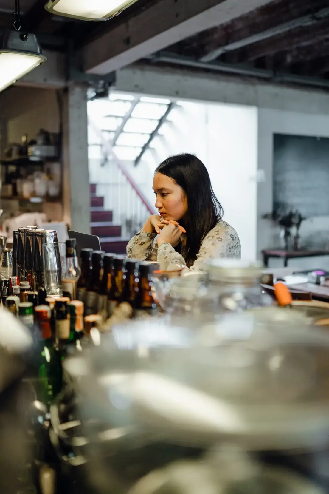 Woman contemplating at a bar counter surrounded by various alcoholic beverages, reflecting on personal drinking habits and relationship with alcohol.