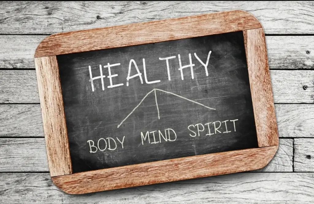 Chalkboard displaying the word "HEALTHY" with arrows pointing to "BODY," "MIND," and "SPIRIT," symbolizing holistic wellness, against a rustic wooden background.